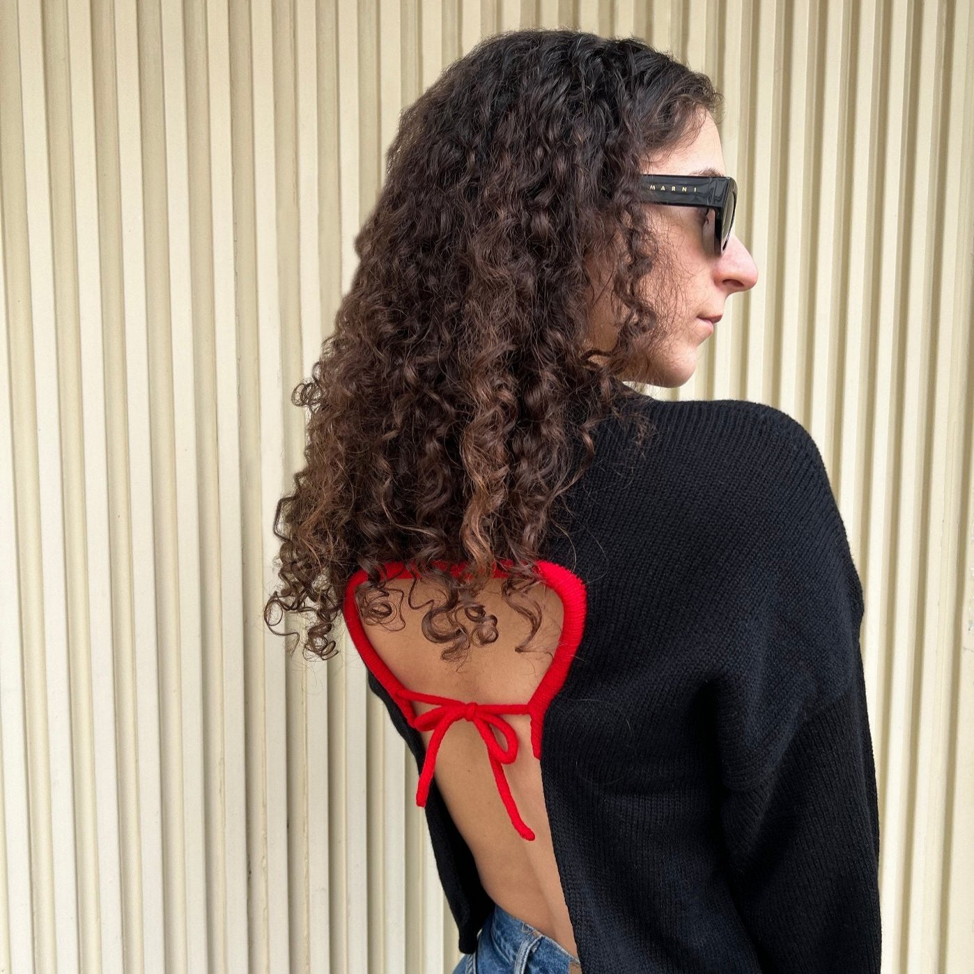 Person wearing a black jacket over a red bikini top and blue jeans, standing against a neutral background.
givéh knitwear handmade by female artisans in iran using only natural and recycled fibres. contemporary knitwear for cool people. crewneck backless sweater in black. 100% merino wool. fall 2025 collection.