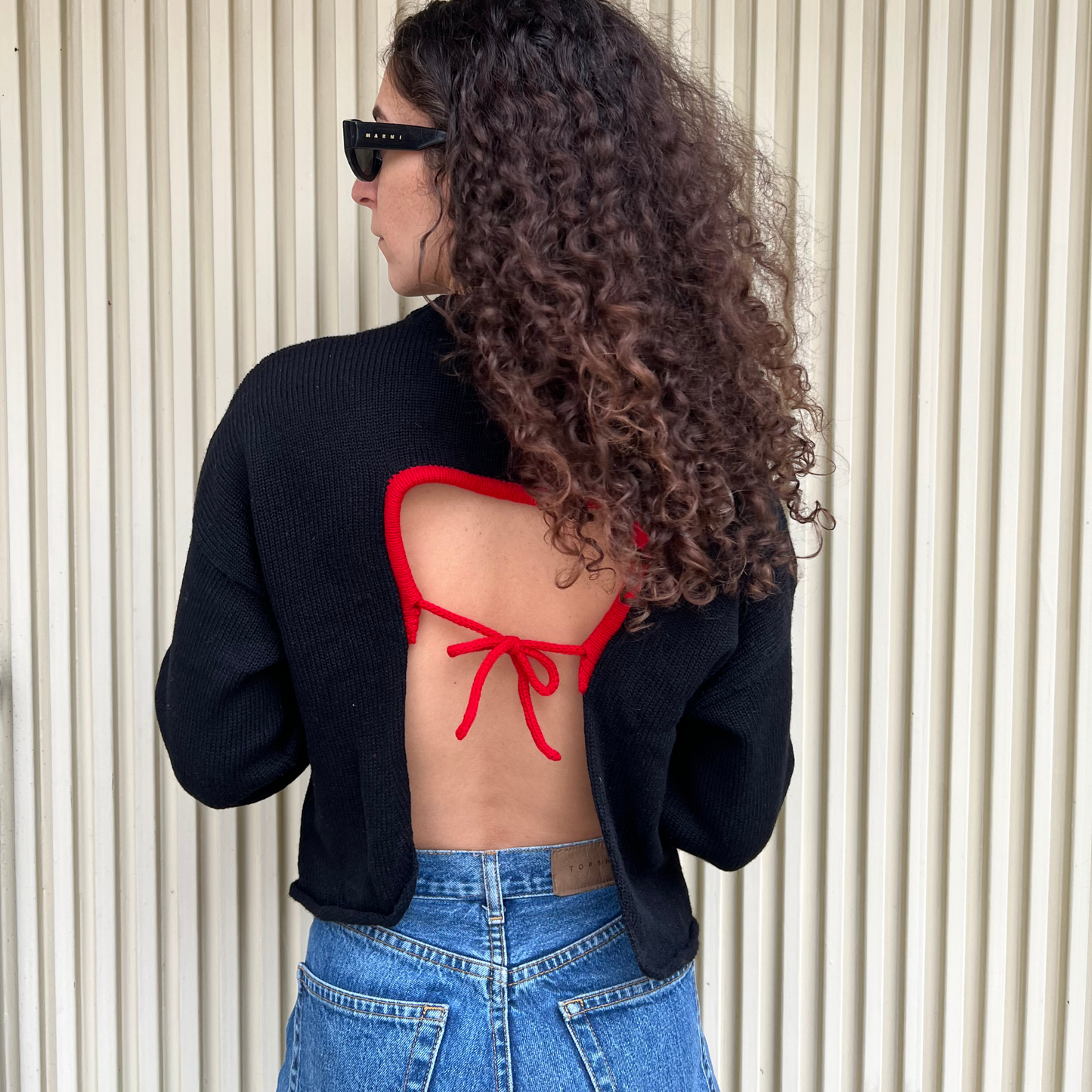 Person wearing a black jacket over a red bikini top and blue jeans, standing against a neutral background.
givéh knitwear handmade by female artisans in iran using only natural and recycled fibres. contemporary knitwear for cool people. crewneck backless sweater in black. 100% merino wool. fall 2025 collection.
