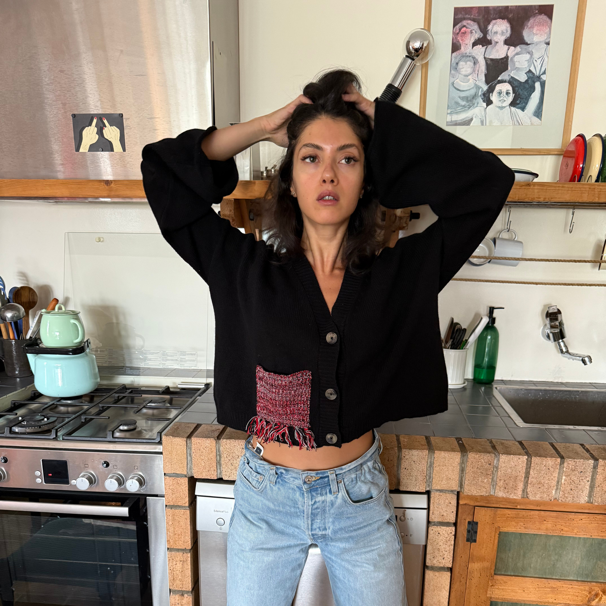 Woman in a kitchen adjusting her hair with a kitchen counter and appliances in the background.
givéh knitwear handmade by female artisans in iran using only natural and recycled fibres. contemporary knitwear for cool people. black cardigan with red/black gradient fringe pocket. 3 natural horn buttons. 100% merino wool. fall 2025 collection.