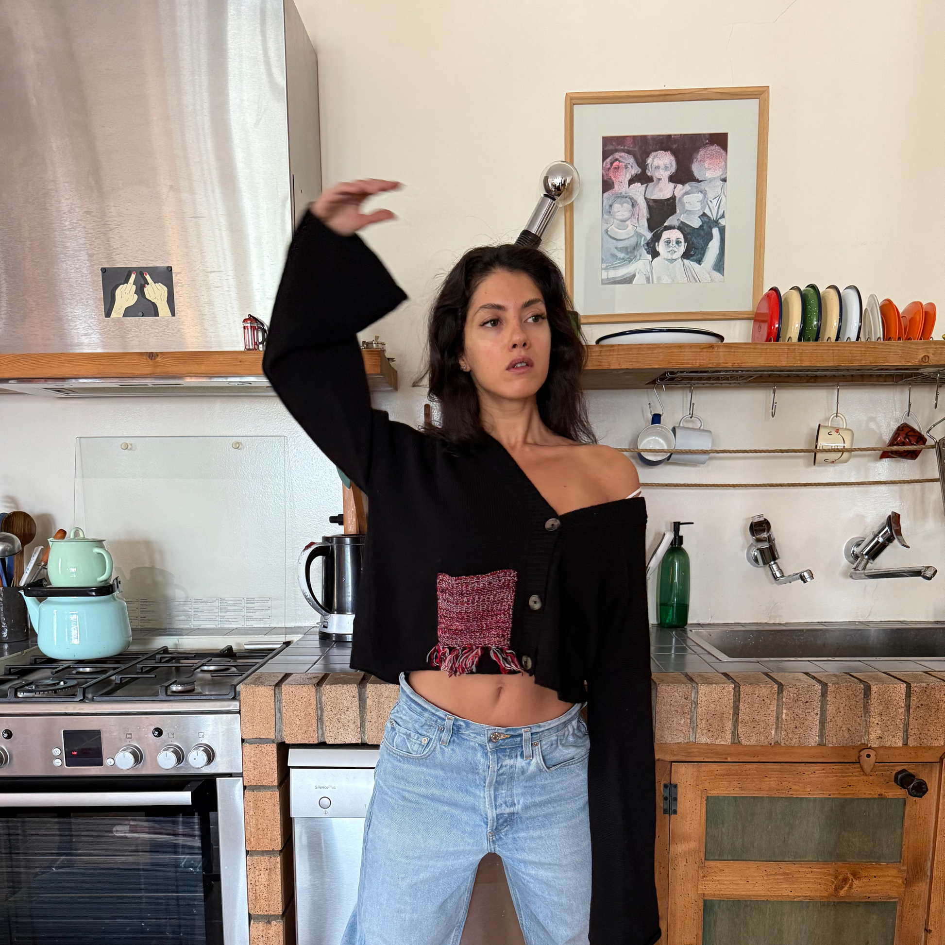 Woman posing in a kitchen with a framed picture on the wall.
givéh knitwear handmade by female artisans in iran using only natural and recycled fibres. contemporary knitwear for cool people. black cardigan with red/black gradient fringe pocket. 3 natural horn buttons. 100% merino wool. fall 2025 collection.