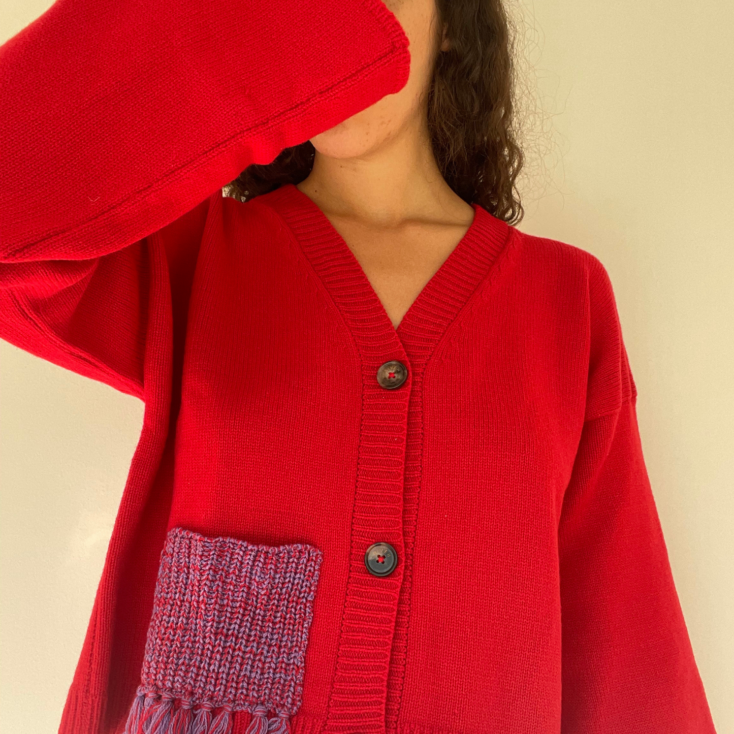 Red cardigan with a pocket on a beige background.
givéh knitwear handmade by female artisans in iran using only natural and recycled fibres. contemporary knitwear for cool people. red cardigan with red/purple gradient fringe pocket. 3 natural horn buttons. 100% merino wool. fall 2025 collection.